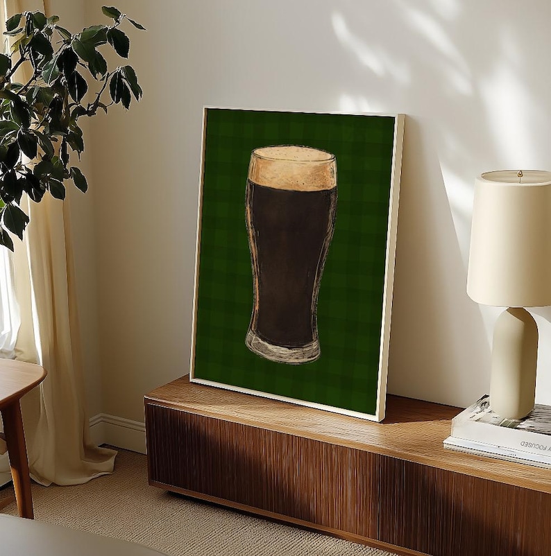May include: A framed print depicting a pint of dark beer with a foamy head. The artwork is set against a green plaid background. The print is displayed on a wooden cabinet.