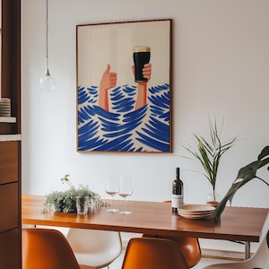 May include: A framed art print depicts two hands emerging from blue waves, one giving a thumbs-up and the other holding a pint of dark beer. The artwork has a brown frame and hangs above a wooden dining table with orange chairs.