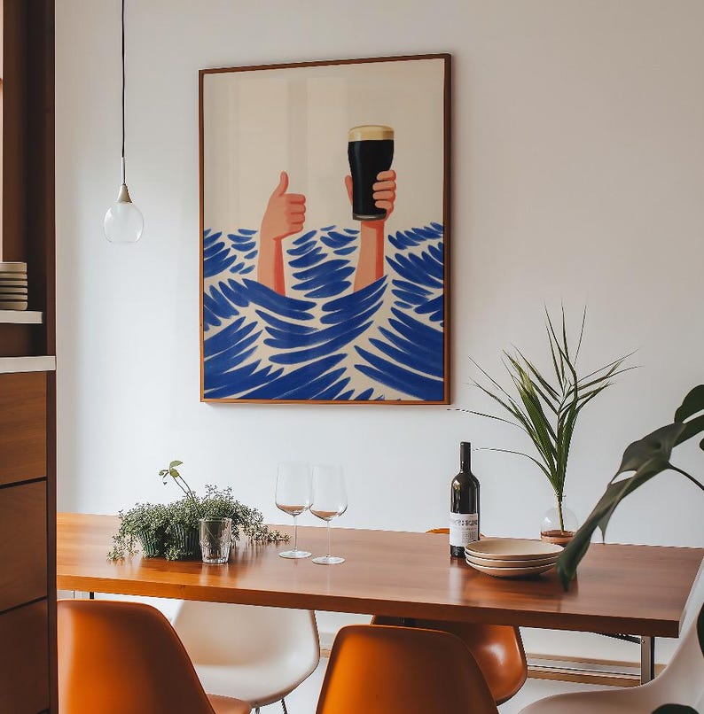 May include: A framed art print depicts two hands emerging from blue waves, one giving a thumbs-up and the other holding a pint of dark beer. The artwork has a brown frame and hangs above a wooden dining table with orange chairs.