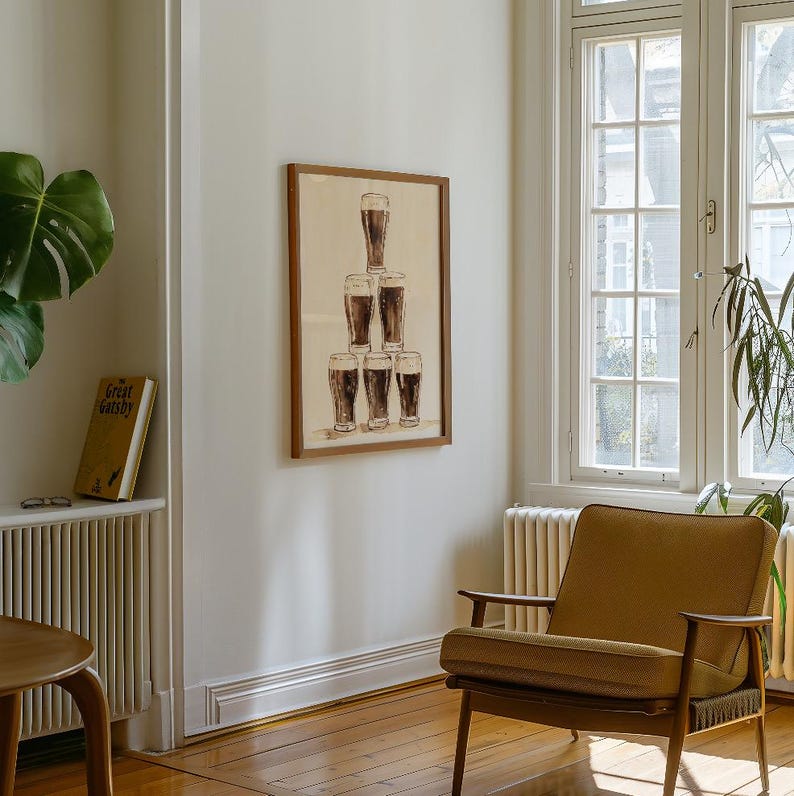 May include: A framed print featuring a pyramid of six glasses filled with a dark beverage. The artwork is displayed on a light-colored wall in a room with a window, a chair, and a plant.