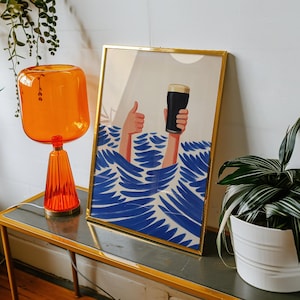 May include: A framed art print with a gold frame depicts a person's hands holding a pint of dark beer above blue waves. One hand gives a thumbs-up. The artwork has a minimalist style and is displayed on a gold-trimmed table.