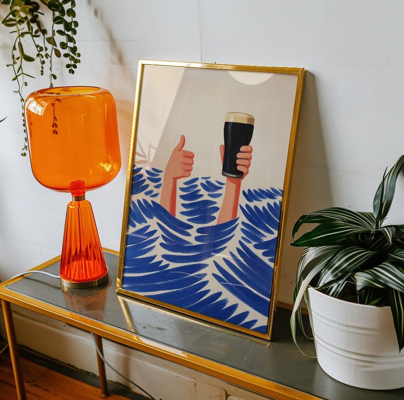 May include: A framed art print with a gold frame depicts a person's hands holding a pint of dark beer above blue waves. One hand gives a thumbs-up. The artwork has a minimalist style and is displayed on a gold-trimmed table.