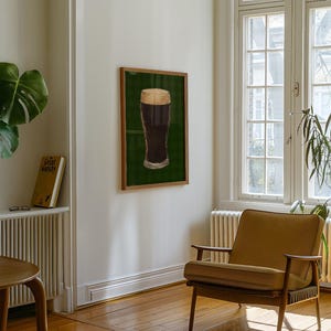 May include: A framed print of a pint of stout with a creamy head, set against a green backdrop. The artwork is in a wooden frame, mounted on a white wall. The room includes a mid-century chair and a window, creating a cozy atmosphere.