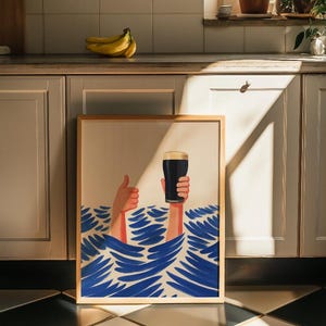 May include: A framed art print featuring a whimsical illustration of two hands emerging from blue waves. One hand gives a thumbs-up, while the other holds a pint of dark beer. The artwork has a light wood frame and is set against a neutral background.