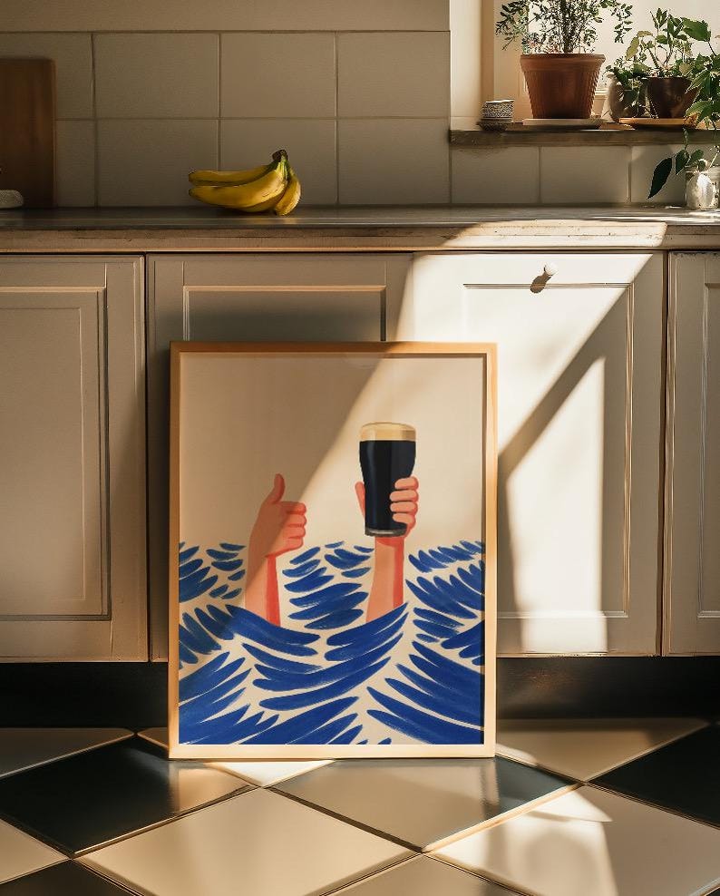 May include: A framed art print featuring a whimsical illustration of two hands emerging from blue waves. One hand gives a thumbs-up, while the other holds a pint of dark beer. The artwork has a light wood frame and is set against a neutral background.