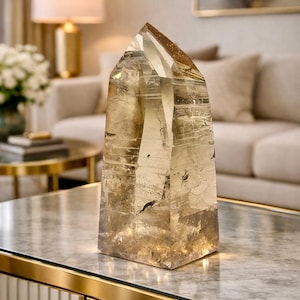 Könnte beinhalten: Ein großer, transparenter Quarz-Kristallpunkt mit goldbraunem Farbton. Der Kristall wird auf einem Tisch mit Marmorplatte und goldenen Zierleisten präsentiert. Die Facetten des Kristalls fangen das Licht ein und erzeugen einen funkelnden Effekt. Thema: Wohnkultur.