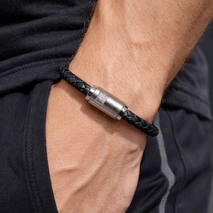 Può includere: Bracciale in pelle intrecciata nera con chiusura argentata. Il bracciale è indossato al polso, mostrando la consistenza della pelle e la chiusura metallica. Un accessorio di moda.