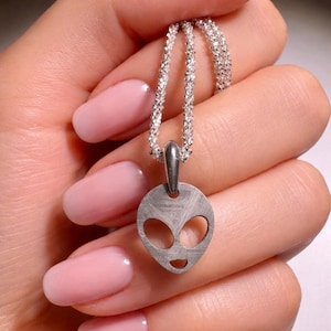 Puede incluir: Collar con colgante plateado con diseño de cara alienígena. El colgante tiene dos cortes ovalados para los ojos y una boca curva. La cadena del collar está hecha de pequeñas cuentas brillantes. Fondo: una mano con esmalte de uñas rosa.