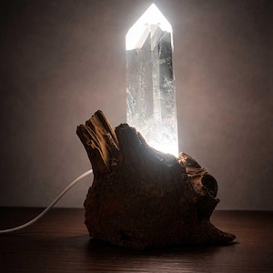 Könnte beinhalten: Eine klare Quarz-Kristall-Lampe, von innen beleuchtet, auf einem verwitterten, braunen Holzsockel. Der Kristall hat eine hohe, rechteckige Form mit einer spitzen Spitze. Ein weißes Netzkabel geht von der Basis aus.