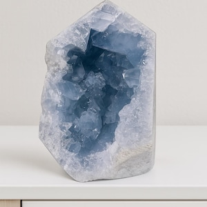 Könnte beinhalten: Eine große, hellblaue Celestin-Kristallgeode. Der Kristall hat eine raue, strukturierte Außenseite und ein tiefblaues Inneres. Die Geode wird auf einer weißen Oberfläche präsentiert, darunter ein weißer und holzfarbener Schrank.