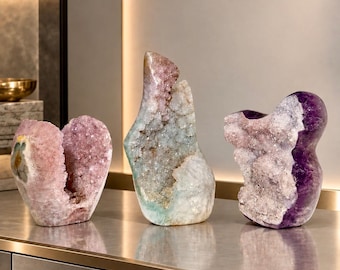 May include: Three distinct crystal geodes displayed on a metallic surface. The left geode is pale pink, the center is a tall, multi-colored formation with light blue and white tones, and the right geode features deep purple and white crystalline structures.
