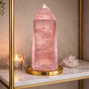 Könnte beinhalten: Ein großer, facettierter Rosenquarz-Kristall-Obelisk auf einem goldfarbenen Metalltablett. Der Kristall hat eine zarte rosa Farbe und steht auf einem Marmorregal mit goldfarbenem Rand. Eine brennende Kerze und ein Cluster aus klaren Quarzkristallen befinden sich ebenfalls auf dem Regal.