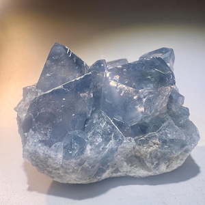 May include: A cluster of light blue celestite crystals. The crystals have a translucent appearance with sharp, angular formations. The base of the cluster is a lighter, more opaque shade of blue. The overall shape is irregular and natural.