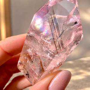 May include: A translucent, pale pink crystal point with a sharp, faceted termination. The stone features internal fractures, natural inclusions, and dark needle-like mineral streaks, with a visible rainbow light refraction on its surface.