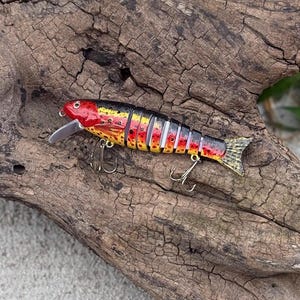 May include: A colorful fishing lure shaped like a fish, with a red head, yellow and black body, and a silver blade. The lure has two treble hooks and a detailed scale pattern. It is designed to attract fish.