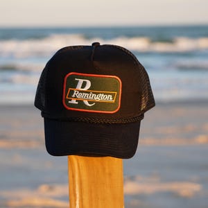 Puede incluir: Una gorra de camionero de malla negra con un parche verde y naranja que dice "Remington" en letras blancas.