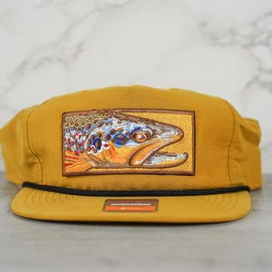 May include: A mustard yellow baseball cap with a brown trout embroidered patch on the front.