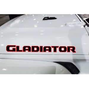 May include: White vehicle hood with the word "GLADIATOR" in bold red letters with a black outline. The hood has a glossy finish and is slightly curved. The image is a close-up of the front of the vehicle.