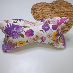 Puede incluir: Una almohada decorativa en forma de lazo con un estampado floral en tonos morado, rosa y amarillo sobre un fondo crema. La almohada se coloca contra un salvamanteles en forma de corazón tejido.
