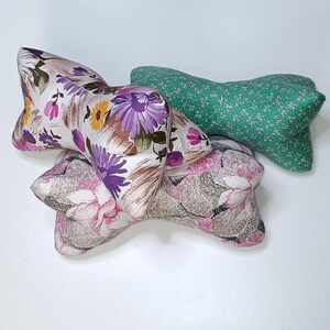 Puede incluir: Tres almohadas decorativas con forma de pajarita. Las almohadas están hechas de tela con estampados florales. Una almohada es verde con flores blancas, otra es blanca con flores moradas y amarillas, y la última es rosa con flores blancas.