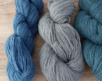 May include: Three skeins of yarn in different shades of blue. The skeins are arranged in a row on a wooden surface.