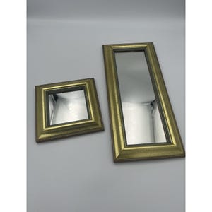 May include: Two mirrors with textured gold frames featuring dark brown inner edges. One mirror is square in shape, while the other is rectangular and elongated. Both mirrors have reflective glass surfaces and are positioned side by side.