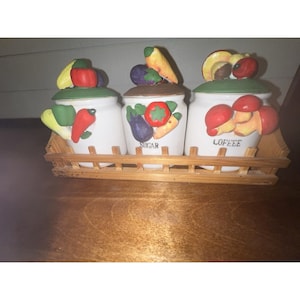 Puede incluir: Un juego de tres tarros de cerámica blanca con tapas y detalles de verduras, colocados en un soporte de madera. El tarro central tiene la etiqueta "SUGAR" y el de la derecha "COFFEE". Cada tapa presenta verduras esculpidas y pintadas.