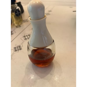 May include: A small, clear glass bottle with a rounded base containing amber-colored liquid. The bottle features a white, bell-shaped cap with a rounded top, creating a distinct silhouette. The design is simple, functional, and compact.