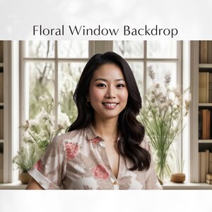 May include: A woman with long dark hair is standing in front of a window with a floral backdrop. She is wearing a light brown blouse with a floral pattern and a white background. The window is framed in white and has a white sill. There are white flowers in a vase on the sill.