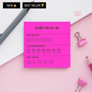 May include: A vibrant pink Daily Check-In notepad with sections for mood, hydration, and sleep. The notepad has a simple design with icons for tracking daily habits. The notepad is on a pink surface with a pen and paper clips.