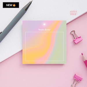 May include: A pastel pink, yellow, and green gradient sticky note with the text "brain dump" in white. The sticky note is on a pink surface with a silver pen, two pink binder clips, and a pink pencil.