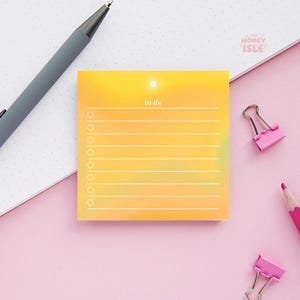 May include: Yellow to-do list notepad with a sun graphic and blank lines for writing. The notepad is on a pink surface with a pink paperclip and pencil.