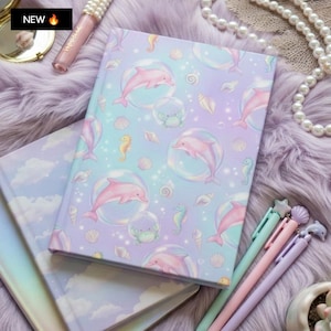May include: A hardcover notebook with a pastel purple and blue gradient cover featuring a repeating pattern of pink dolphins inside bubbles, small crabs, seahorses, and seashells. The notebook is accompanied by three pens with star, shell, and dolphin toppers.