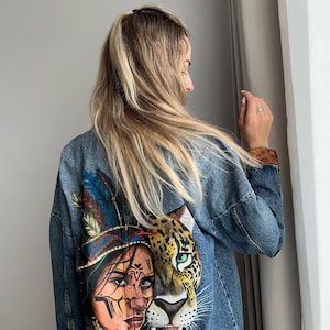 Könnte beinhalten: Jeansjacke mit handgemaltem Design. Das Kunstwerk zeigt eine indianische Frau und das Gesicht eines Leoparden, mit detaillierter Verwendung von Farben wie Blau, Braun und Grün. Die Jacke hat einen klassischen Jeans-Stil.