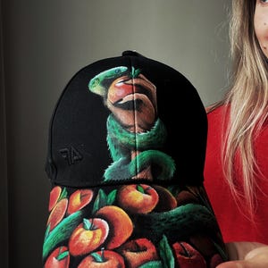 Pode incluir: Boné de beisebol preto com um design pintado à mão. O boné apresenta uma cobra verde enrolada em torno de uma maçã vermelha, com uma aba adornada com maçãs e folhas verdes. O boné tem uma aba curva e uma coroa estruturada.