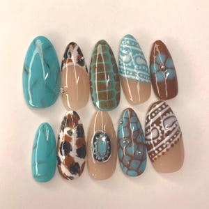 Może przedstawiać: Zestaw dziesięciu sztucznych paznokci o różnych wzorach. Paznokcie mają kolory turkusowy, brązowy i beżowy z wzorami takimi jak marmur, zwierzęcy nadruk i geometryczne wzory. Niektóre paznokcie mają małe srebrne zdobienia.
