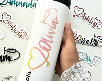 May include: White Owala water bottle with a black lid and handle. The bottle features the name "Olivia" in a gradient pink and yellow font with a stethoscope design. Several name stickers with stethoscope designs are scattered around the bottle.