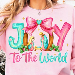 May include: A light pink sweatshirt with a colorful graphic that says "JOY To The World." The design features a pink bow, a nativity scene, and the letters "J" and "Y" in turquoise. The sweatshirt is a festive holiday item.