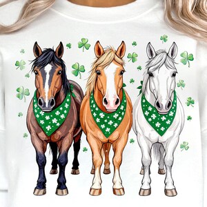 Puede incluir: Sudadera blanca con un gráfico de tres caballos con bandanas verdes con patrones de trébol. Los caballos son marrón, blanco y naranja. El fondo está cubierto con pequeños patrones de trébol.