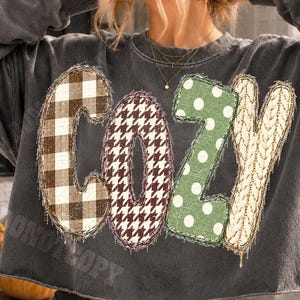 May include: A charcoal gray cropped sweatshirt featuring the word COZY printed in large, textured letters. Each letter has a distinct pattern: the C is brown plaid, the O is brown houndstooth, the Z is green with white polka dots, and the Y is cream cable knit.
