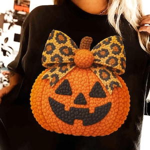 May include: Black sweatshirt featuring a large, orange pumpkin design with a smiling face and a leopard print bow. The pumpkin is the focal point, perfect for Halloween or fall. The sweatshirt is a comfortable, casual style.