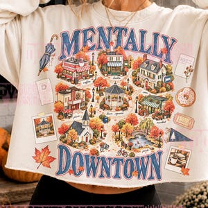 May include: A cream-colored cropped sweatshirt featuring a colorful graphic print of a town scene with autumn trees, a gazebo, and various buildings. The text 'MENTALLY DOWNTOWN' is printed in large blue letters above and below the central illustration.