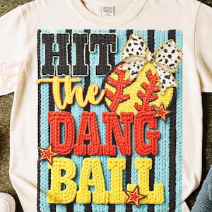 May include: Cream-coloured t-shirt with a graphic that says "Hit The Dang Ball." The graphic features a baseball with a bow, and stars. The text is in black, red, and yellow. The background is blue and black stripes.
