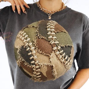 Puede incluir: Camiseta gris oscuro con un diseño circular que representa una pelota de patchwork. El diseño de la pelota incorpora varias telas en tonos tierra, incluyendo verdes, marrones y beige, cosidas con hilo color crema. La camiseta tiene un aspecto vintage y desgastado.