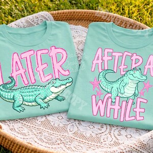 May include: Two light teal t-shirts with alligator graphics. One shirt says "LATER" in pink glitter letters above a green alligator. The other says "AFTER A WHILE" with a similar alligator and pink stars.