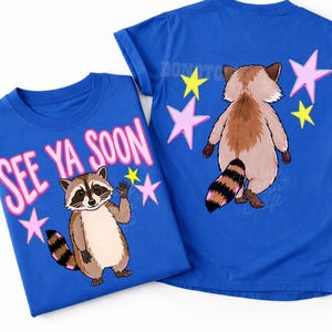 K&ouml;nnte beinhalten: Zwei blaue T-Shirts mit Waschb&auml;r-Grafiken. Eines zeigt einen winkenden Waschb&auml;ren mit dem Text "SEE YA SOON" in Rosa. Das andere zeigt die R&uuml;ckseite eines Waschb&auml;ren mit Sternen. Beide Shirts haben einen Rundhalsausschnitt.