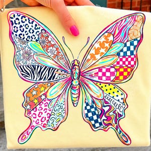 May include: A pale yellow t-shirt featuring a large, colorful butterfly graphic. The butterfly wings are divided into segments with various patterns, including leopard print, floral motifs, zebra stripes, stars, checkerboard, and polka dots.
