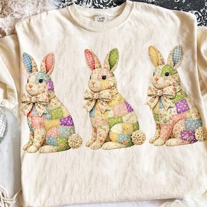 May include: Cream-colored t-shirt featuring three seated bunny rabbits. Each bunny has a patchwork design in pastel colors, a bow tie, and a fluffy tail. The bunnies' ears are patterned with polka dots and gingham.