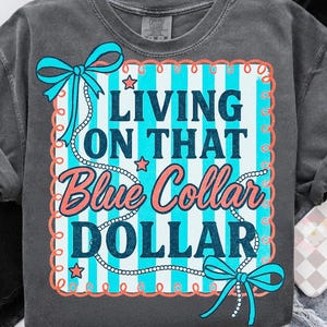 May include: Dark grey t-shirt with the text "LIVING ON THAT Blue Collar DOLLAR" in a blue and coral design. The design includes a blue and white striped background, stars, and decorative bows.
