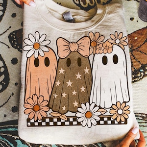 May include: Cream-colored sweatshirt featuring a whimsical design of three ghost figures adorned with flowers and bows. The ghosts are in shades of peach, brown, and white, with star and daisy accents. A black and white checkered border completes the design.
